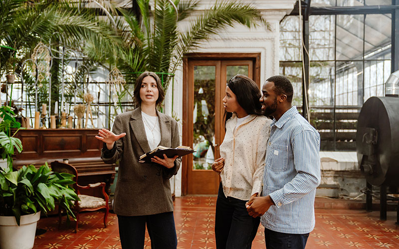 A woman and a man stand and hold hands while listening to a female organizer standing next to them and holding a clipboard while consulting